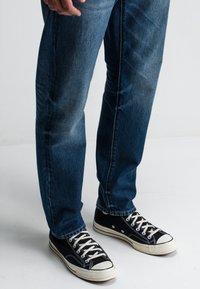 Dunkelblaue Jeans mit entspannter Passform, kombiniert mit schwarzen Canvas-Sneakern mit weißen Gummisohlen und Schnürsenkeln.