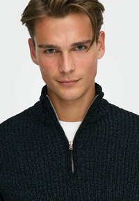 Dunkelblauer Strickpullover mit Stehkragen und Reißverschluss, strukturiertem Muster und dezentem Glanz. Über einem weißen Hemd getragen.