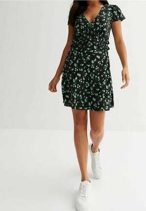 Femme portant une robe noire à motif floral à manches courtes et des baskets blanches, marchant devant un fond blanc uni.