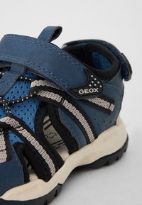 Blå och svart sandal i mocka och meshmaterial, utrustad med en kardborreband, lätt sula och geometrisk detaljering på sidan.