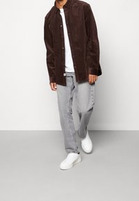 Hnědá manšestrová košile, pod ní bílého tričko, šedé džíny a bílé tenisky. Košile má knoflíky a volný střih.