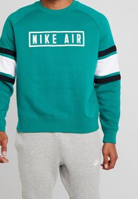 Tealfärgad sweatshirt med vita, svarta och tealfärgade ärmstriper, har en central vit ruta med "NIKE AIR" text, ribbad halsringning och ärmmuddar.