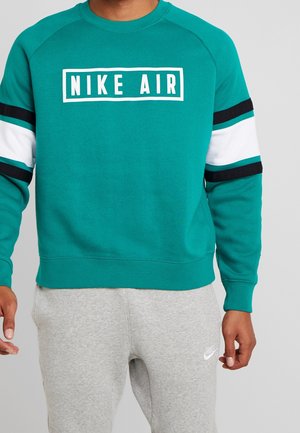 Sudadera azul petróleo con rayas en blanco, negro y azul petróleo en las mangas, presenta un cuadro central blanco con el texto "NIKE AIR", y cuellos y puños acanalados.