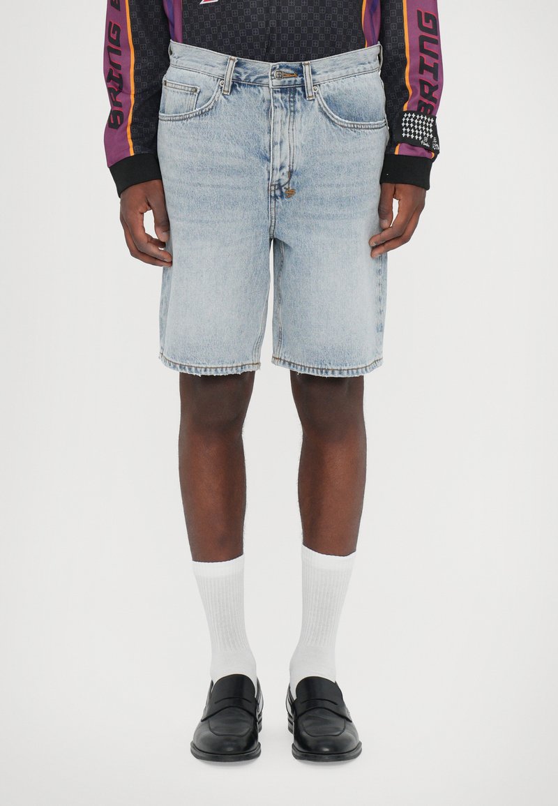 Shorts din denim de culoare albastră deschisă, cu o croială clasică, finisate cu un tiv brut. Asortate cu șosete albe cu nervuri și mocasini negri.