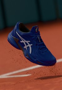 Baskets de sport bleues avec un dessus texturé, logo blanc et accents contrastants, suspendues au-dessus d'une piste sablonneuse avec un flou de mouvement.