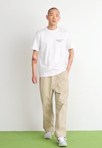 Tricou alb din bumbac cu un grafic mic, asortat cu pantaloni bej lejeri și adidași gri deschis; fundal simplu.