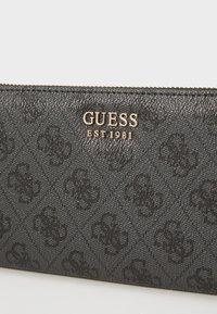 Zwarte leren portefeuille met een herhaald logo-patroon, goudkleurige letters "GUESS EST 1981" en een ritsluiting langs de bovenrand.