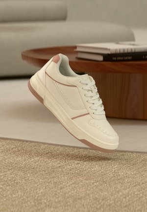 Sneaker color crema con accenti marrone chiaro, lacci bianchi, posizionata vicino a un tavolino in legno e un divano beige in un soggiorno moderno.