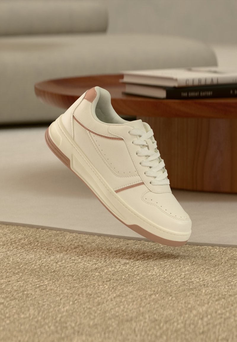 Sneaker color crema con accenti marrone chiaro, lacci bianchi, posizionata vicino a un tavolino in legno e un divano beige in un soggiorno moderno.