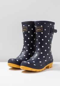 Laivastonsininen pilkullinen kumisaapas sateeseen, jossa on teksturoitu keltainen pohja; sisältää solkidetalin ja logotarran sivulla.