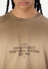 Brun bomulds T-shirt med rund halsudskæring og et falmet tryk: "THE CORE PROJECT NEW VISUAL SYSTEM EST. 1990" i mørk skrift.