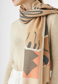 Femme portant un pull beige et un pantalon blanc avec une écharpe beige présentant des motifs abstraits marron, noir et orange enroulée autour de son cou.