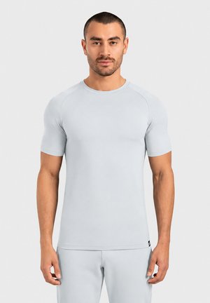 Hombre con cabello corto y oscuro y barba, vestido con una camiseta de manga corta ajustada gris claro y pantalones a juego, de pie frente a un fondo liso.
