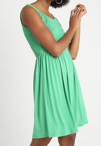 Grön ärmlös klänning med smockad bodice och full kjol. Slät tyg med mjuk fall, knälång och figurnära i midjan.