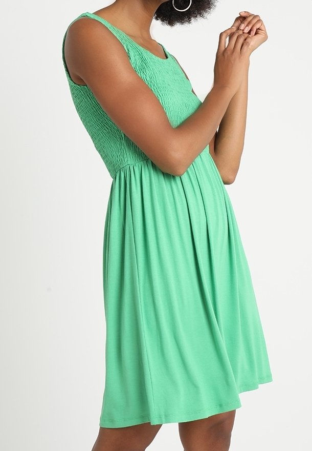 Grön ärmlös klänning med smockad bodice och full kjol. Slät tyg med mjuk fall, knälång och figurnära i midjan.