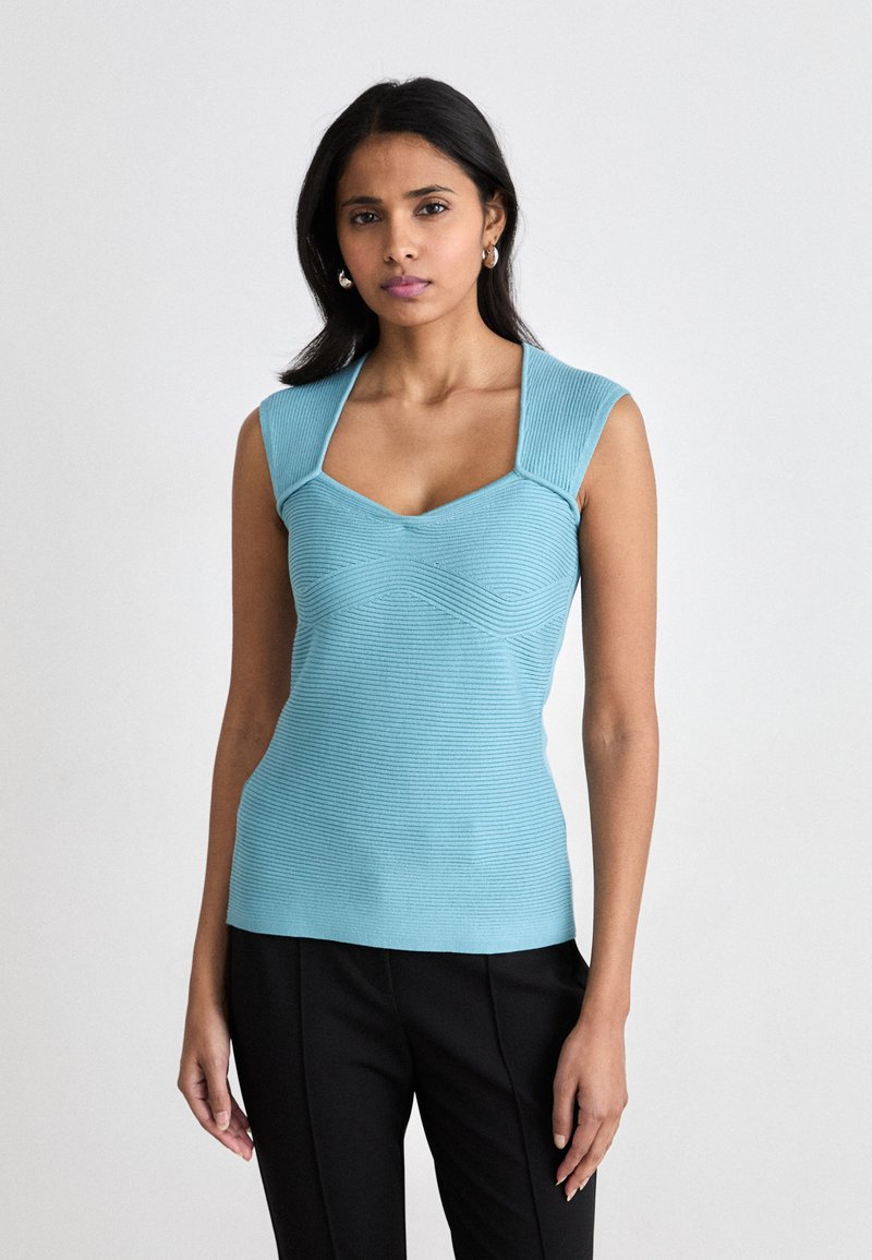 Top senza maniche di colore azzurro chiaro con scollatura quadrata e un sottile motivo a onde. Realizzato in tessuto elasticizzato, abbinato a pantaloni neri.