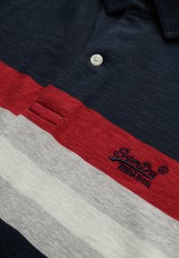 Camiseta polo de base azul marino con rayas horizontales en rojo, gris y blanco. Presenta un logo bordado y detalles de botones en el collar.
