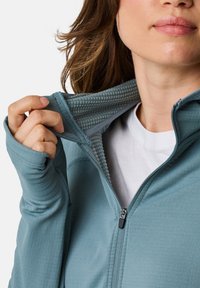 Blue zip-up jacket with a textured inner collar, visible zip closure, and thumbholes at the sleeves, worn over a white shirt.