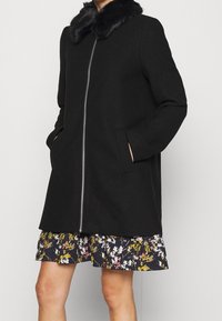 Cappotto nero con collo in pelliccia sintetica, cerniera frontale e tasche laterali. Il design è a taglio dritto. Indossato sopra un vestito a motivo floreale.