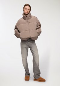 Giacca imbottita trapuntata color taupe con colletto alto, indossata sopra jeans grigi sbiaditi e scarpe slip-on in suede marroni.