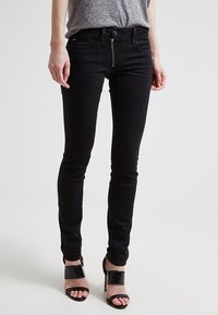Black skinny jeans made of denim, featuring a zip fly, front pockets, and a smooth texture. Paired with strappy black heels.