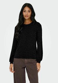 Svart genser med lange ermer, teksturert mønster, rund hals og tettsittende mansjetter, kombinert med brune bukser.