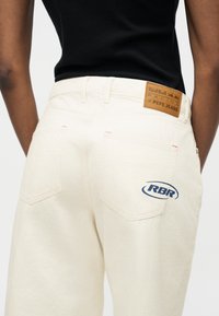 Άτομο που φορά τζιν σε κρεμ απόχρωση με δερμάτινο ταμπελάκι Red Bull Racing x Pepe Jeans και το λογότυπο RBR στην πίσω τσέπη, συνδυασμένο με μαύρο τοπ.