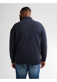 Marinblå stickad tröja med hög ribbad krage, texturerat mönster, sänkta axlar och ribbade muddar och fåll. Lämplig för vardagsbruk.