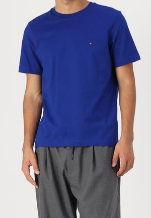 Mężczyzna ubrany w prosty królewsko-niebieski t-shirt z krótkim rękawem, z małym logo flagi na piersi, oraz luźne szare spodnie.