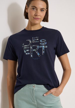 Femme portant un T-shirt bleu marine avec le mot "DESERT" en lettres à motif et un pantalon vert clair, main dans la poche.
