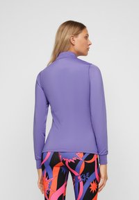 Langärmliges lila Oberteil mit Stehkragen. Glattes Material und taillierte Passform, kombiniert mit bunten, gemusterten Leggings darunter.