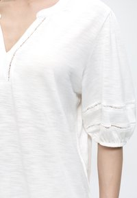 Blouse blanche avec encolure en V et manches courtes bouffantes présentant des détails en point ajouré horizontal.