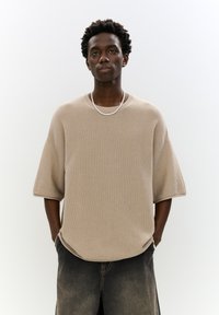 Bežová štruktúrovaná pletená oversized tričko s okrúhlym výstrihom a krátkymi rukávmi, v spojení s tmavo sivými džínsovými šortkami.