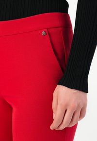 Detailní záběr na osobu v červených přiléhavých kalhotách s pinem Hugo a černém žebrovaném topu s dlouhým rukávem, jejíž ruka je uvolněná při těle.