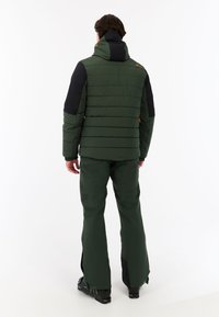 Giacca isolante verde e nera con cappuccio, caratterizzata da un design trapuntato, dettagli arancioni e tasche laterali con zip; abbinata a pantaloni coordinati.