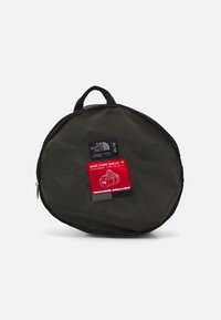 Sac de voyage en mesh vert foncé, de forme ronde, avec des accents noirs, doté d'une poignée supérieure et d'une étiquette rouge incluant les détails du produit.