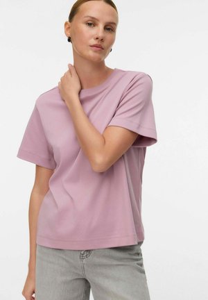 Lyserød kortærmet t-shirt med rund halsudskæring, lavet af blødt stof. Modellen har grå jeans på og rører ved sin hals. Neutral baggrund.