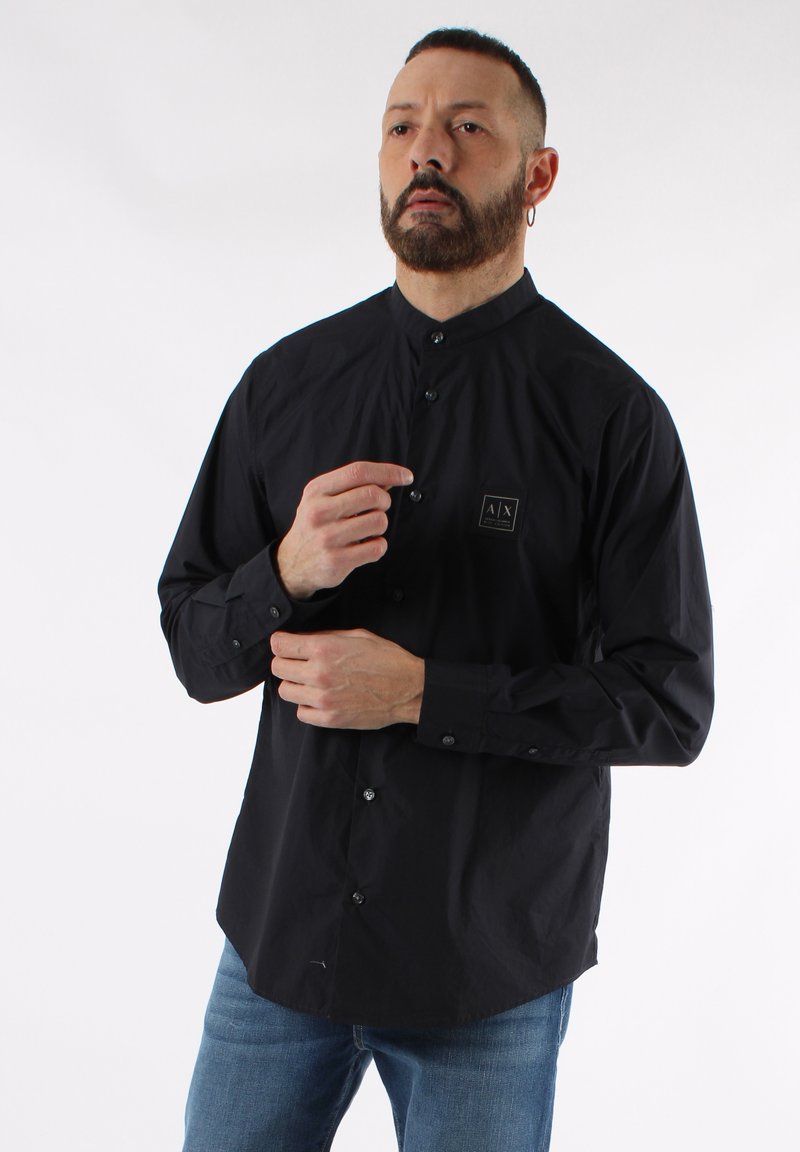 Camicia nera con colletto, maniche lunghe, chiusura con bottoni e piccolo logo ricamato sul petto. Tessuto liscio, vestibilità casual, abbinata a jeans blu.