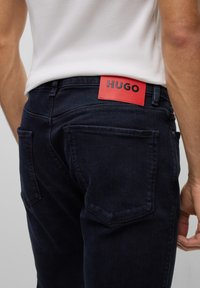 Tmavě modré džíny s úzkým střihem, které mají na zadním pasu viditelnou červenou koženou nášivku s nápisem "HUGO" a viditelné zadní kapsy.