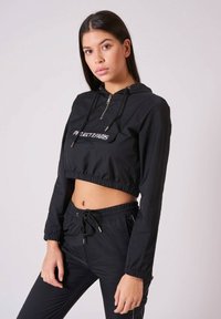 Schwarze, cropped Jacke mit Kapuze, Reißverschluss und elastischem Saum. Verfügt über ein vorderes Logo-Patch und passende schwarze Hosen mit elastischem Bund.