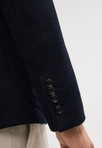 Manica di un maglione lavorato a maglia blu navy con texture a coste, caratterizzata da cinque bottoni neri lungo il polsino.