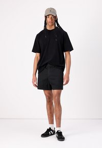 Černé oversize tričko, černé šortky a černé tenisky Adidas s bílými akcenty. Šedá kšiltovka dotváří ležérní outfit.