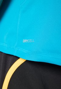 Tirkizna atletska majica s teksturiranim materijalom, sadrži srebrni logo "DRYCELL". Crne kratke hlače istaknute žutom akcentnom linijom.