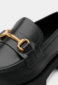 Mocassin en cuir noir avec une finition lisse, doté d'un détail en métal doré sur le dessus et d'un petit talon épais.