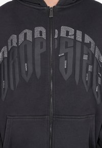 Sort zip-hoodie med stort "EUROPA TEGN" tekst hen over brystet, omkranset af små sølvnitter og en frontkangaroo-lomme.