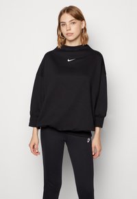 Juoda striukė su aukštu apykakle ir nusileidusiomis petnešomis. Baltas Nike logotipas ant krūtinės. Pora su juodomis legging'ais, kuriuose yra mažas logotipas.
