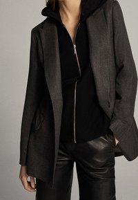 Grijze visgraat blazer met schouderklappen over een zwarte zip-up hoodie, gecombineerd met zwarte leren broek. Geweven stof en op maat gemaakte pasvorm.