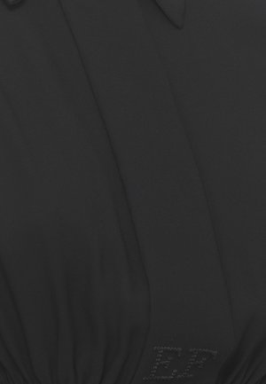 Primo piano di tessuto nero con lettere "EF" cucite e cucitura visibile con arricciature vicino al fondo, suggerendo un dettaglio del capo.