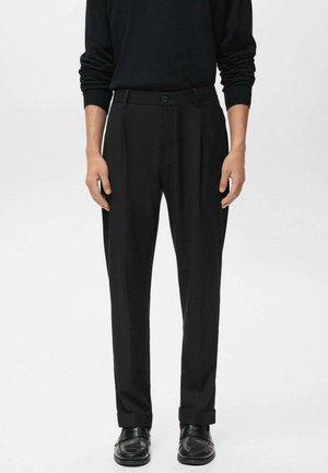 Man wearing black pleated trousers with cuffed hems, a black long-sleeve top, and black leather shoes, standing against a plain background.