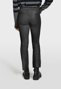 Crne hlače od umjetne kože s prilagođenim krojem, elastičnim pojasom i suptilnim stražnjim džepovima. Noge imaju blagi flare i glatku teksturu.